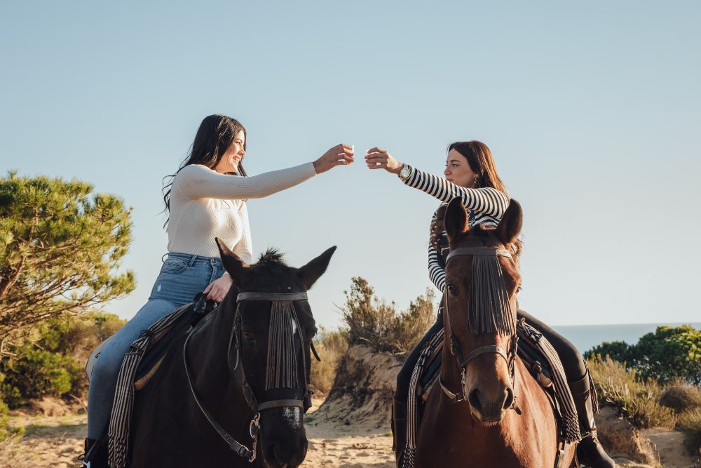 Young happy girlfriends clinking glasses with drinks while riding horses in countryside against cloudless sky and ocean during summer trip